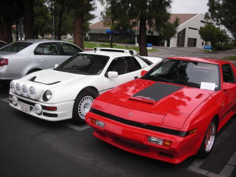 1998 Ford Crown Victoria - A white rally car with prominent fog lights is parked next to a red sports car, with a silver sedan visible in the background.