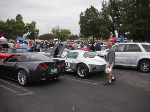 1998 Ford Crown Victoria - A dark gray Chevrolet Corvette is parked at an outdoor car meet, with other vehicles and people in the background.