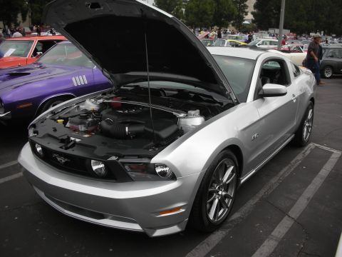1998 Ford Crown Victoria - A silver Ford Mustang GT 5.0 is parked with its hood open, revealing the engine bay, next to a purple Plymouth Barracuda.