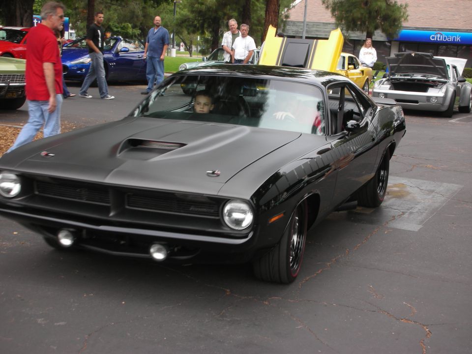 Black 1970 Plymouth Barracuda with a child in the driver's seat. Other cars and people are visible in the background