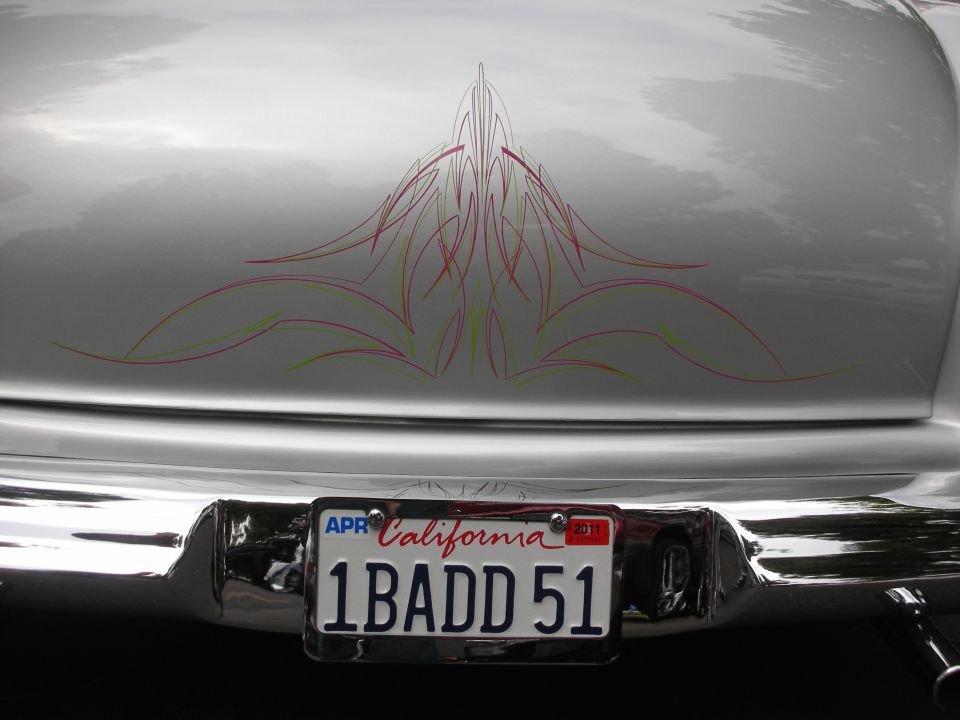 Rear of a silver classic car with red and yellow pinstripes above a California license plate reading 1BADD51
