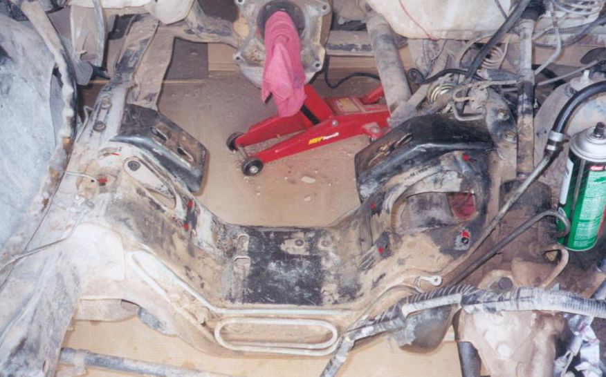 Engine bay of 1987 Ford Bronco with engine removed, dirty crossmember and red floor jack below