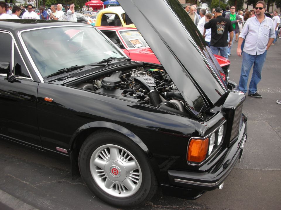 Black Bentley coupe, hood open showing engine. Red car behind it. People standing around