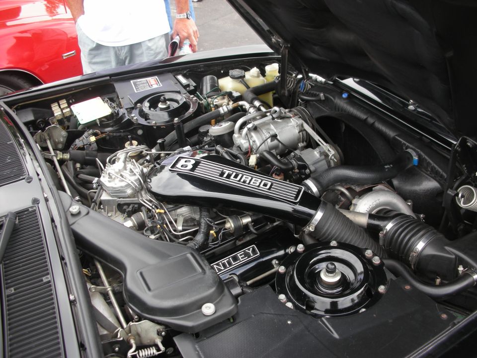 Engine bay of a Bentley with 'B TURBO' and 'BENTLEY' visible on the engine cover. Another car is partially visible to the left