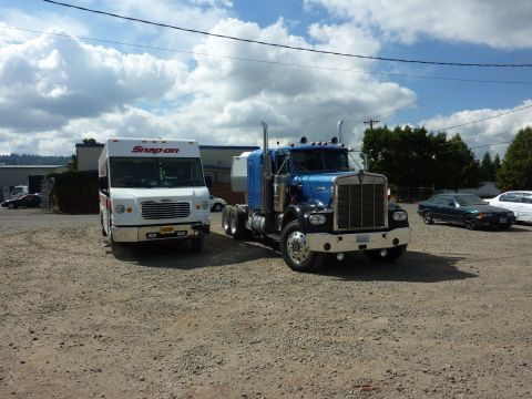 1981 Kenworth W900A - Two trucks and a few cars are parked in a gravel lot under a partly cloudy sky.