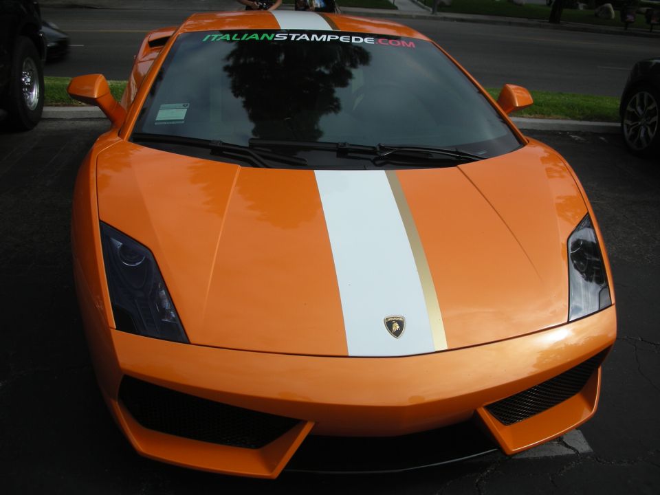 Orange sports car with white and gold stripes on hood, "ITALIANSTAMPEDE.COM" on windshield. Another car is partially visible on the left