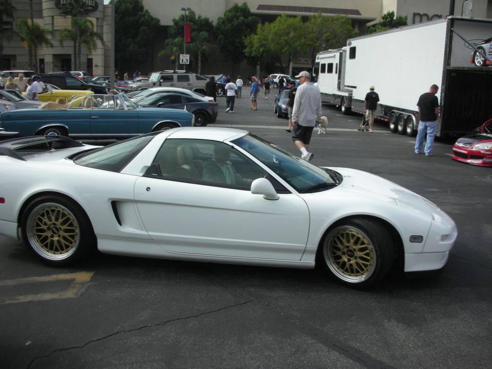 White sports car with gold BBS wheels parked in a lot. Other cars and a trailer are visible