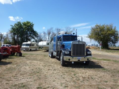 1981 Kenworth W900A - A blue semi-truck is parked in a dry, grassy lot with an RV and a red tractor nearby under a clear blue sky.