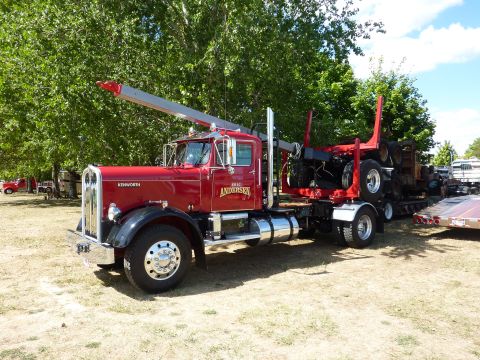 1981 Kenworth W900A - A red Kenworth logging truck is parked with a log trailer attached, with other vehicles visible in the background.