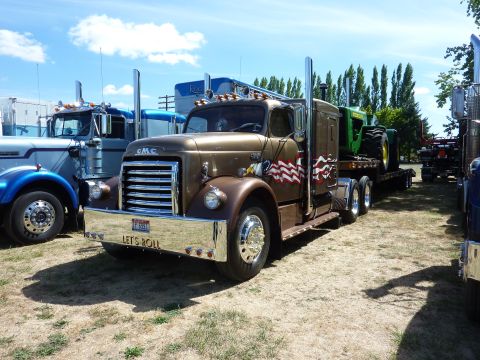 1981 Kenworth W900A - A classic GMC truck is parked next to other vehicles, carrying a tractor on its trailer under a sunny sky.