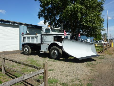 1981 Kenworth W900A - A light gray dump truck equipped with a large snow plow is parked in front of a building.