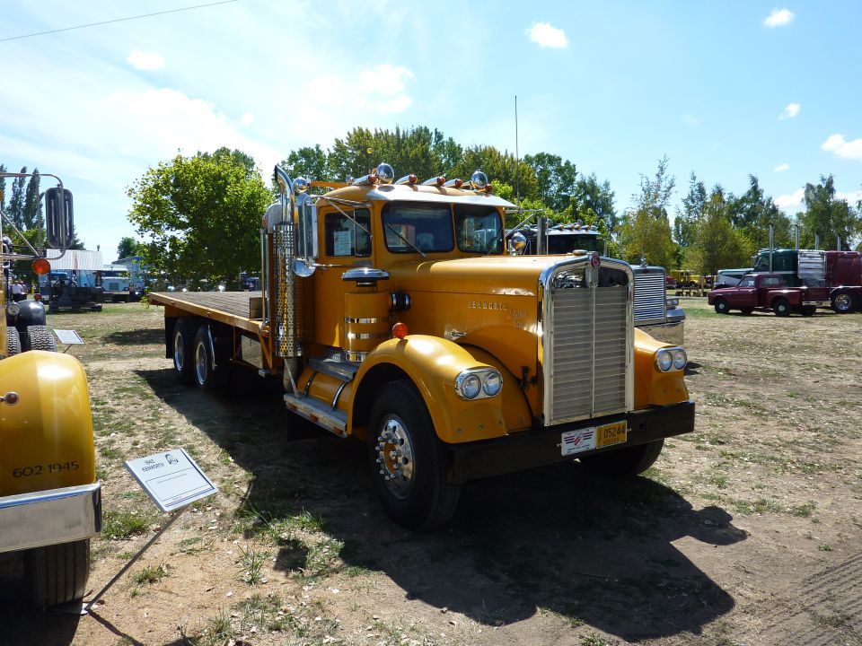 Yellow flatbed semi-truck with chrome grille and stacks parked outdoors. Other trucks and a red pickup are visible in the background