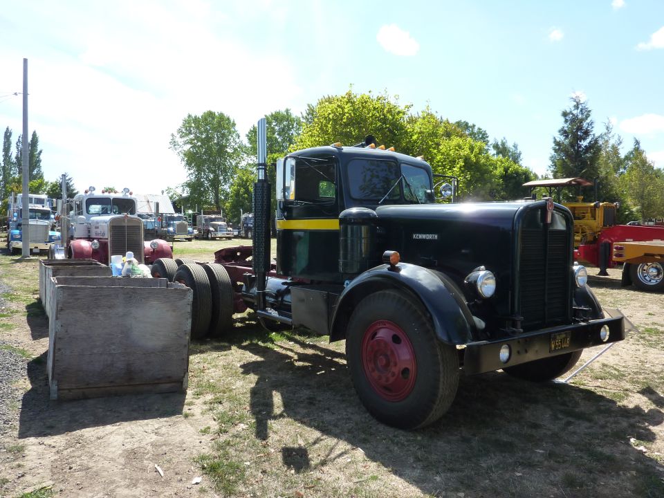Black Kenworth semi-truck with yellow stripe on cab, parked outdoors with other trucks in background. Wooden bin in foreground