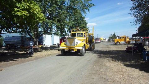 1981 Kenworth W900A - A yellow and white semi-truck drives down a dirt road, with other vehicles and tents visible in the background.