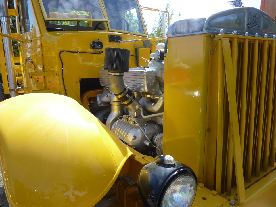 Yellow truck engine with exposed supercharger and chrome grille. Front headlight visible