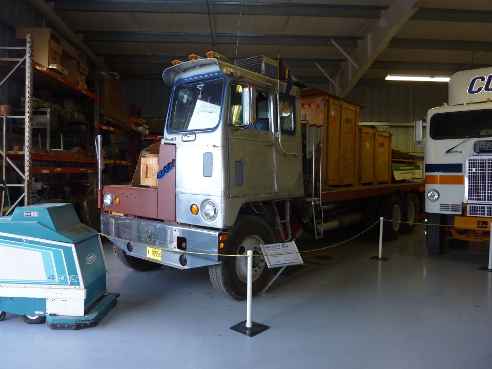 Truck with a flatbed trailer, parked indoors next to another truck and a floor scrubber