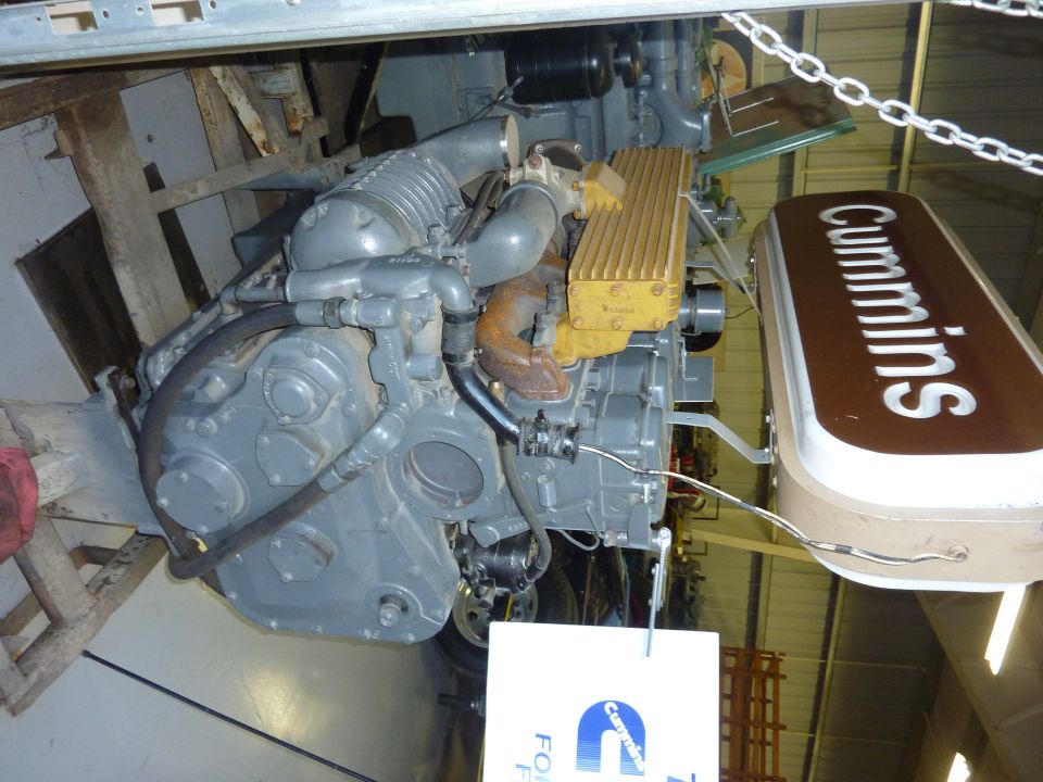 Gray Cummins engine with yellow valve cover and brown sign. Mounted on a wooden stand