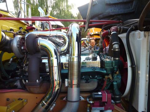1981 Kenworth W900A - Close-up view of a polished chrome turbocharger and exhaust system on a diesel engine, with other trucks visible in the background.