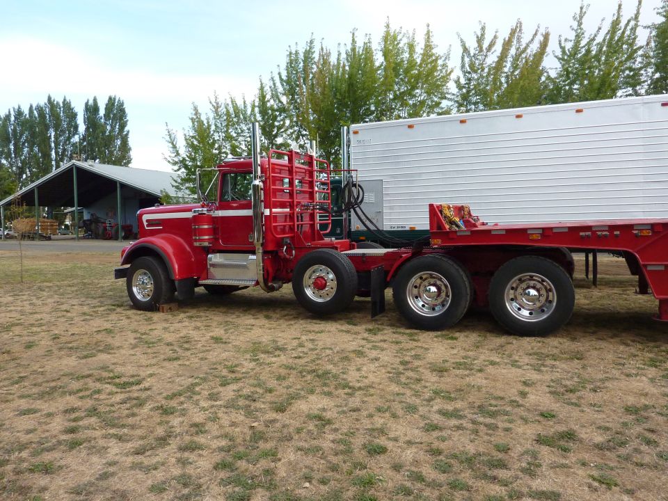 red semi-truck hooked to a white trailer, parked on dry grass near a barn