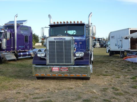 1981 Kenworth W900A - A blue Freightliner truck is parked outdoors, with other trucks and a tent in the background.
