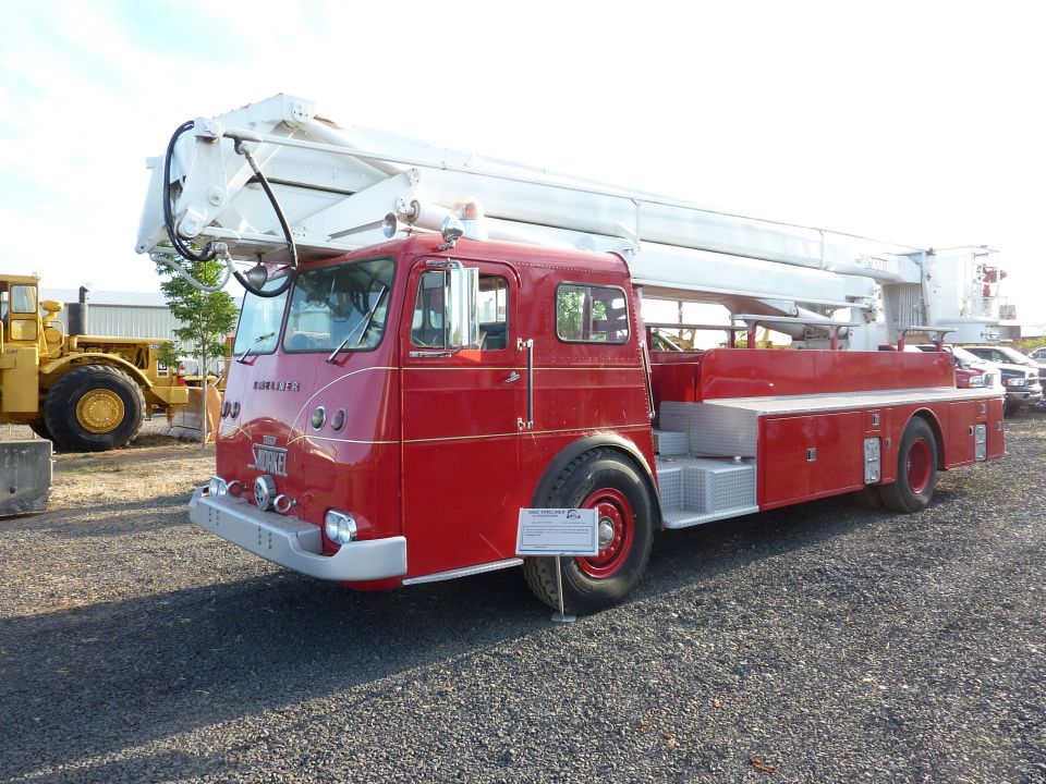 red fire truck with a white ladder extended, parked on gravel. Yellow construction vehicle in background