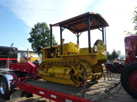 1981 Kenworth W900A - A yellow crawler tractor with a wooden canopy is secured to a trailer, with other vehicles visible in the background.
