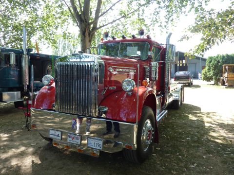 1981 Kenworth W900A - A red semi-truck is parked outdoors with trees and buildings in the background. Another truck is visible to the left.