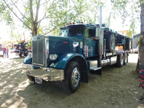 1981 Kenworth W900A - A blue Peterbilt 379 truck is parked on a dirt lot under trees, with another truck visible in the background.
