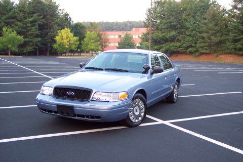 2001 Ford Crown Victoria - A blue 2001 Ford Crown Victoria is parked in a lot with trees and a building in the background.