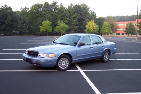 2001 Ford Crown Victoria - A blue 2001 Ford Crown Victoria sedan is parked in a nearly empty parking lot with trees and a building in the background.