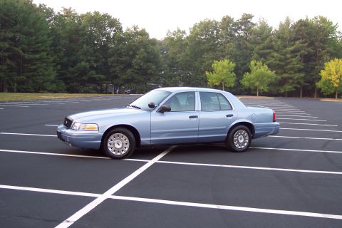 2001 Ford Crown Victoria - This light blue 2001 Ford Crown Victoria sedan is parked in a nearly empty lot with trees in the background.