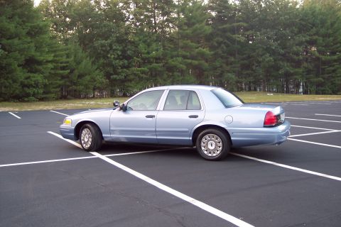 2001 Ford Crown Victoria - A light blue 2001 Ford Crown Victoria sedan is parked in an empty lot with trees in the background.