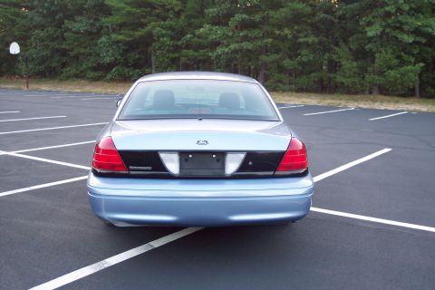 2001 Ford Crown Victoria - The rear of a light blue 2001 Ford Crown Victoria is shown parked in an empty lot with trees in the background.