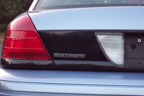 2001 Ford Crown Victoria - The rear of a light blue 2001 Ford Crown Victoria Police Interceptor is visible, showing the taillight and 'Police Interceptor' badging.
