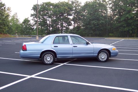2001 Ford Crown Victoria - A light blue 2001 Ford Crown Victoria is parked in a nearly empty lot with trees in the background.