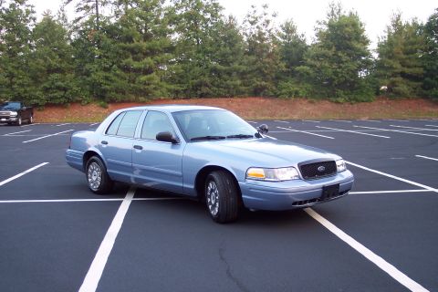 2001 Ford Crown Victoria - A blue 2001 Ford Crown Victoria police car is parked in a lot, with a dark pickup truck visible in the distance.
