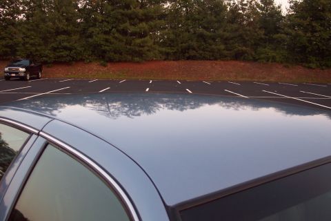 2001 Ford Crown Victoria - A blue sedan's roof reflects trees and sky, with a black pickup truck parked in a lot behind it.