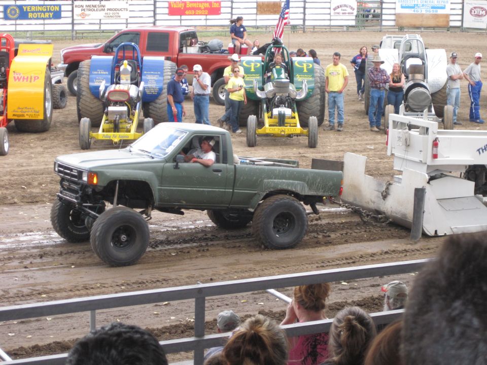 modified green pickup truck pulling a sled on a dirt track, other tractors and vehicles in background