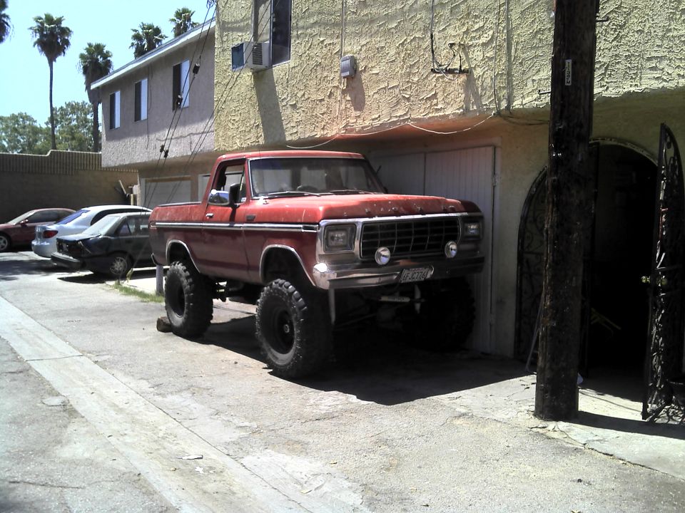 Red 1978 Ford Bronco pickup truck with lifted suspension and large tires parked in front of a building