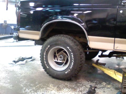 1990 Ford Bronco - Close side view of the rear quarter of a black Ford truck/SUV with a tan lower two-tone stripe, sitting in a shop on a lift with a BFGoodri…