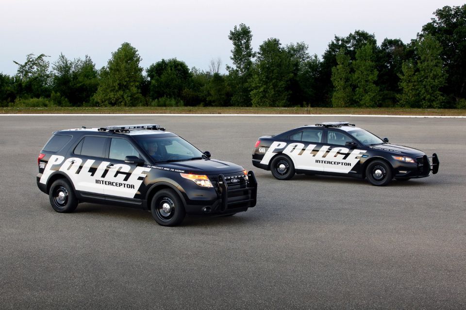 Two black and white police interceptor vehicles, one SUV and one sedan, parked on asphalt with trees in the background