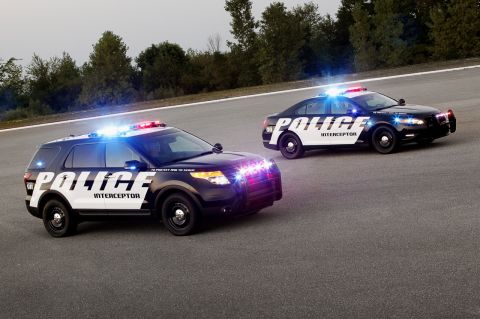 1993 Ford Taurus - Two police interceptor SUVs drive on a paved surface with trees in the background.