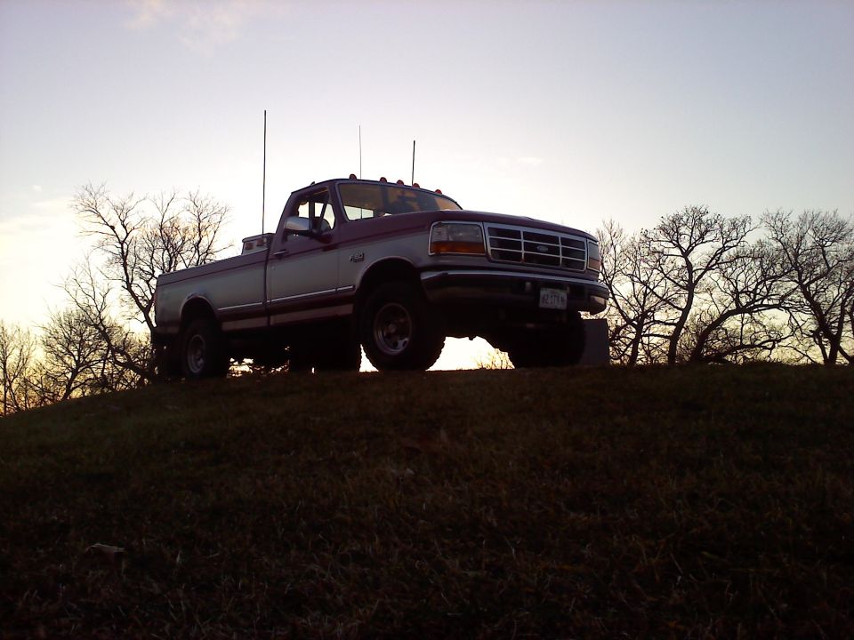 Two-tone pickup truck parked on a grassy hill against a hazy sky. It has dual antennas and a lifted suspension