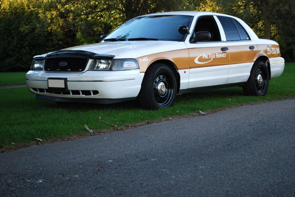 White and tan 1998 Ford Crown Victoria taxi with black wheels, parked on grass next to a road