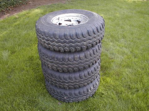1980 Ford Bronco - Four off-road tires with aggressive tread patterns are stacked on white rims outdoors.