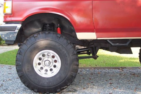 1990 Ford Bronco - Close-up side view of a red SUV, highlighting its large off-road tire and suspension system. The vehicle appears to be a 1990 Ford Bronco.