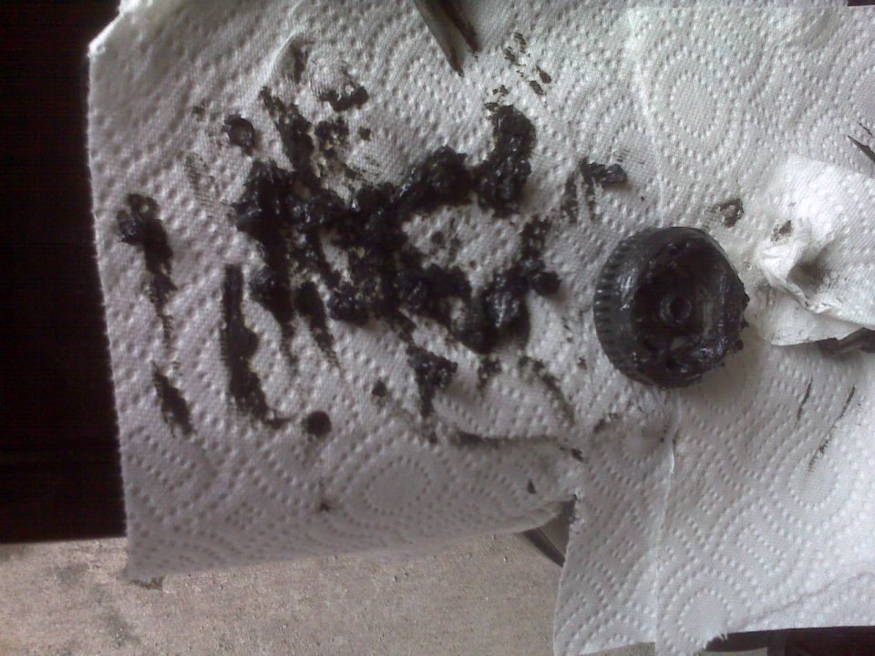 Sludge-covered round black cap or part on a white paper towel with black grease smears