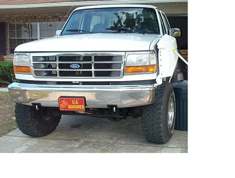 1995 Ford Bronco - Front end of a lifted white full-size Ford truck shown up close, with a chrome grille and blue Ford oval, amber turn signals, a chrome bump…