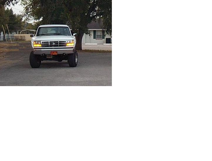 Front view of lifted Ford pickup on dirt lot, headlights on, chrome grille, large tires