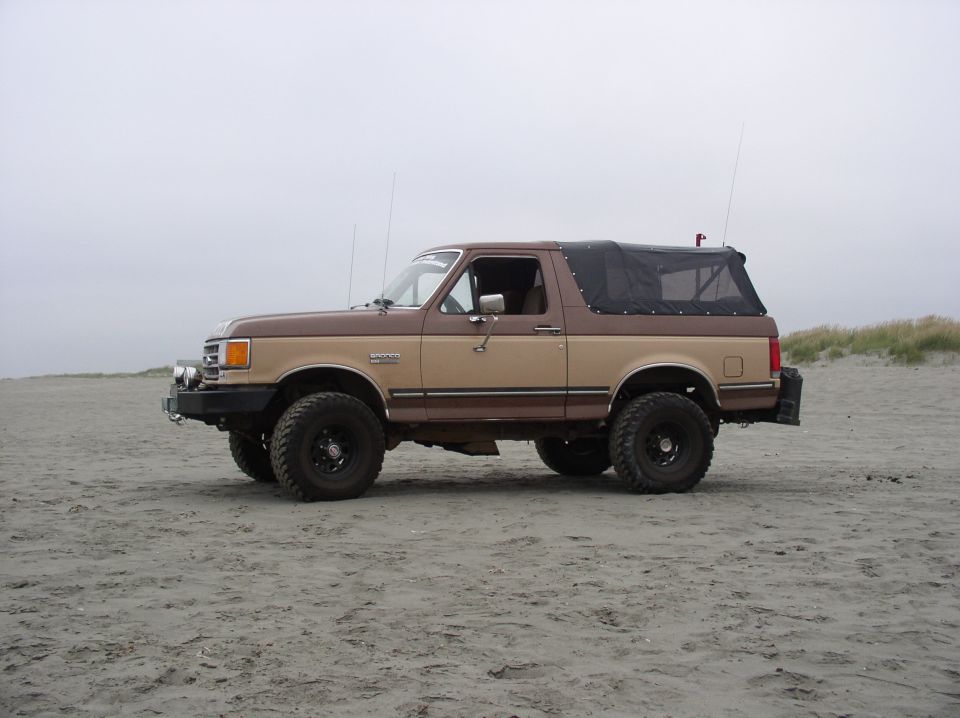 Two-tone brown and tan lifted Ford Bronco with black soft top parked on a sandy beach, side view